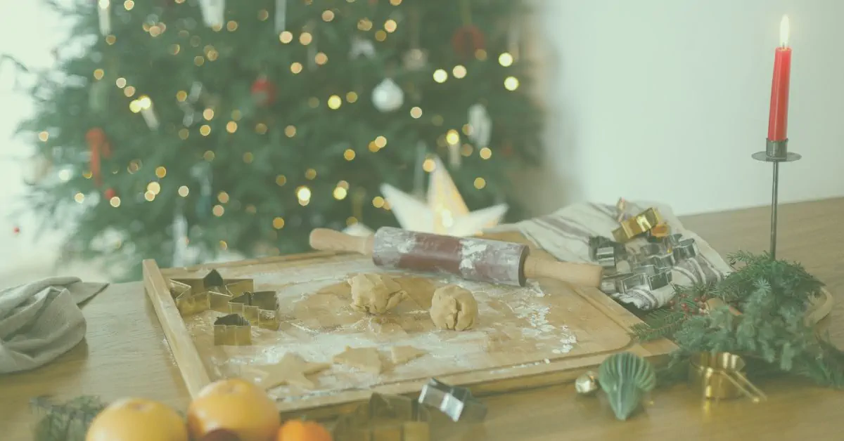 Cosy festive baking scene: wooden board with rolling pin, flour, cookie cutters and dough balls in front of a softly lit Christmas tree with bokeh lights and a glowing red candle.